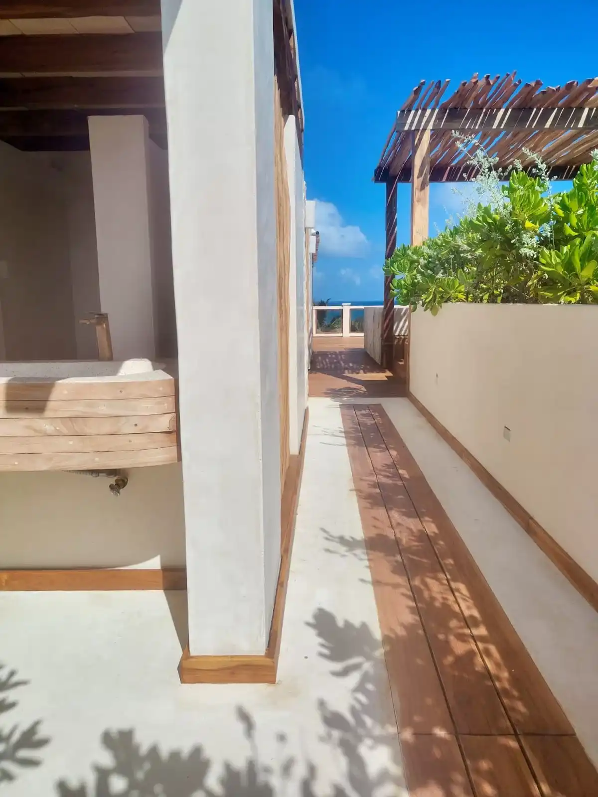 Paso del roof top entre un lavabo de piedra y una pérgola de madera, con duela, jardinera y el mar al fondo.