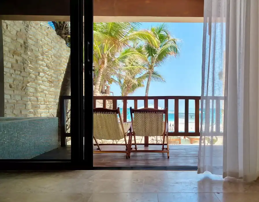 Vista desde el interior de una habitación hacia el balcón: cortinas, dos sillas de lona, palmeras y el mar.
