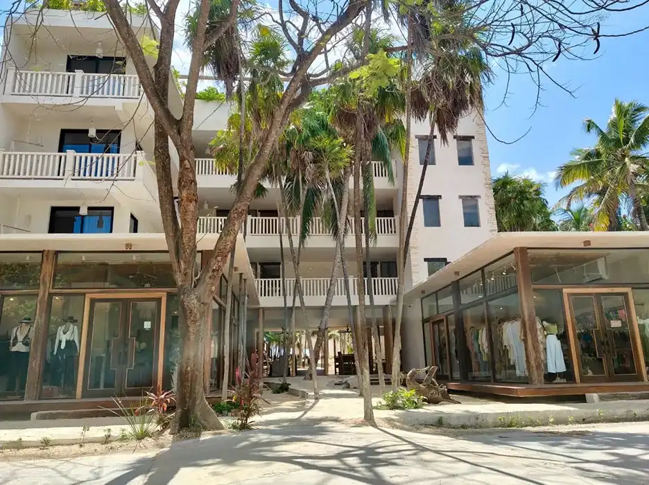 Fachada del hotel desde la calle, con balcones blancos entre los árboles y locales de tiendas en la planta baja.
