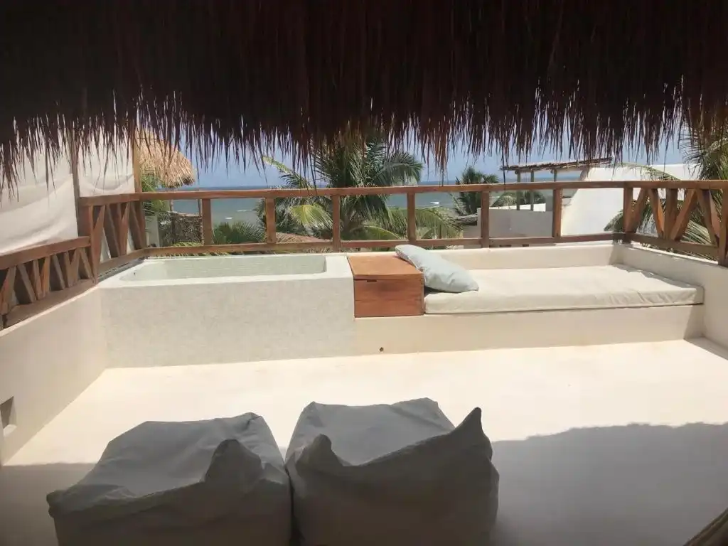 Terraza de azotea con un camastro blanco y cojines, bajo el alero de palma.
