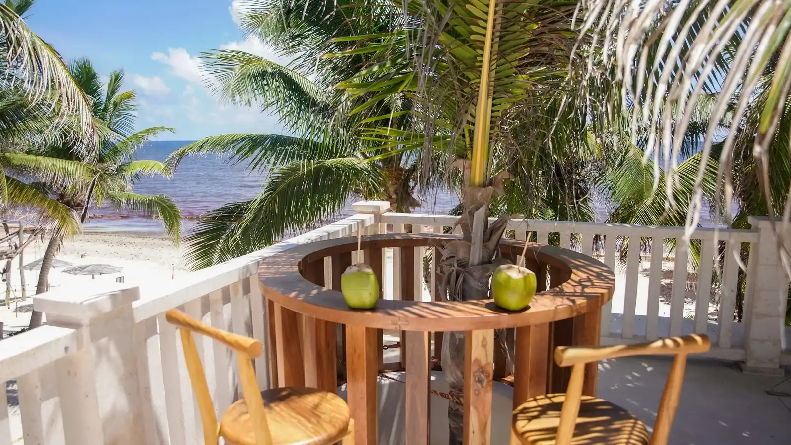 Mesa alta de madera con cocos verdes y banquetas, en una terraza frente a las palmeras y el mar.