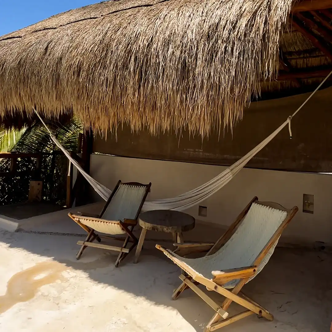 Palapa sobre la arena con una hamaca y dos sillas de tijera de madera.