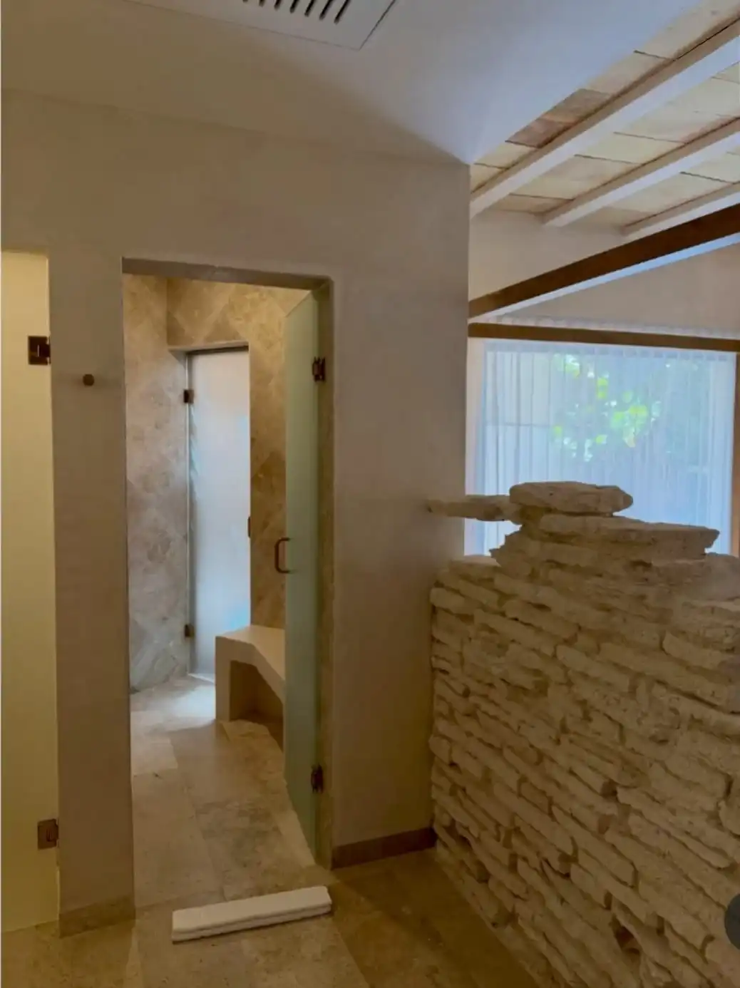 Paso al baño: muro de piedra apilada, puerta de cristal y, detrás, la regadera con su banca de travertino.
