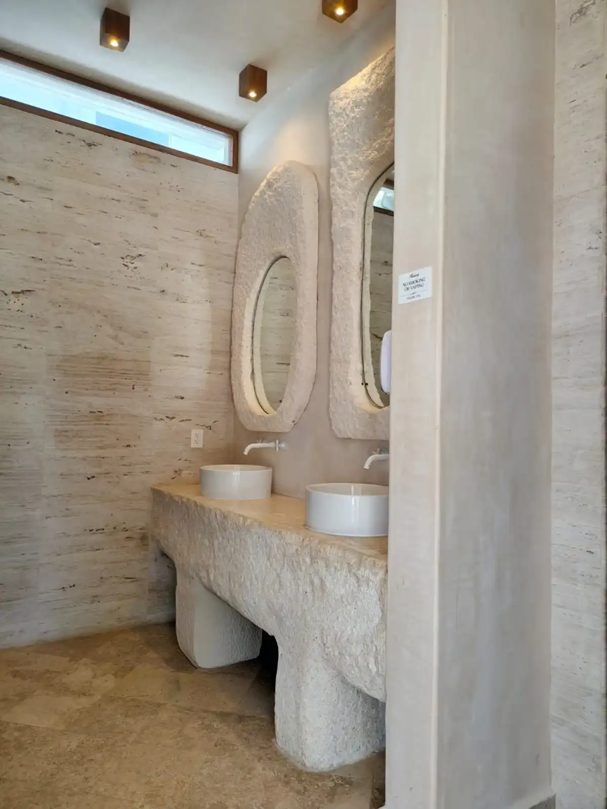 Baño de travertino con dos espejos ovalados enmarcados en piedra y dos lavabos redondos sobre una encimera labrada.