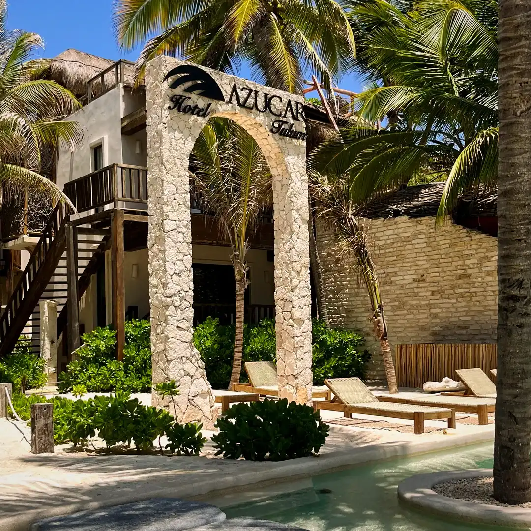 El arco de piedra con el rótulo del hotel, la escalera y los camastros sobre la arena.
