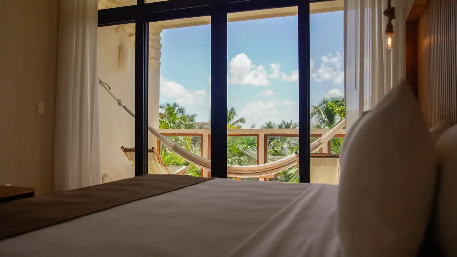 Cama frente a un ventanal, con una hamaca en el balcón y el mar entre las palmas.