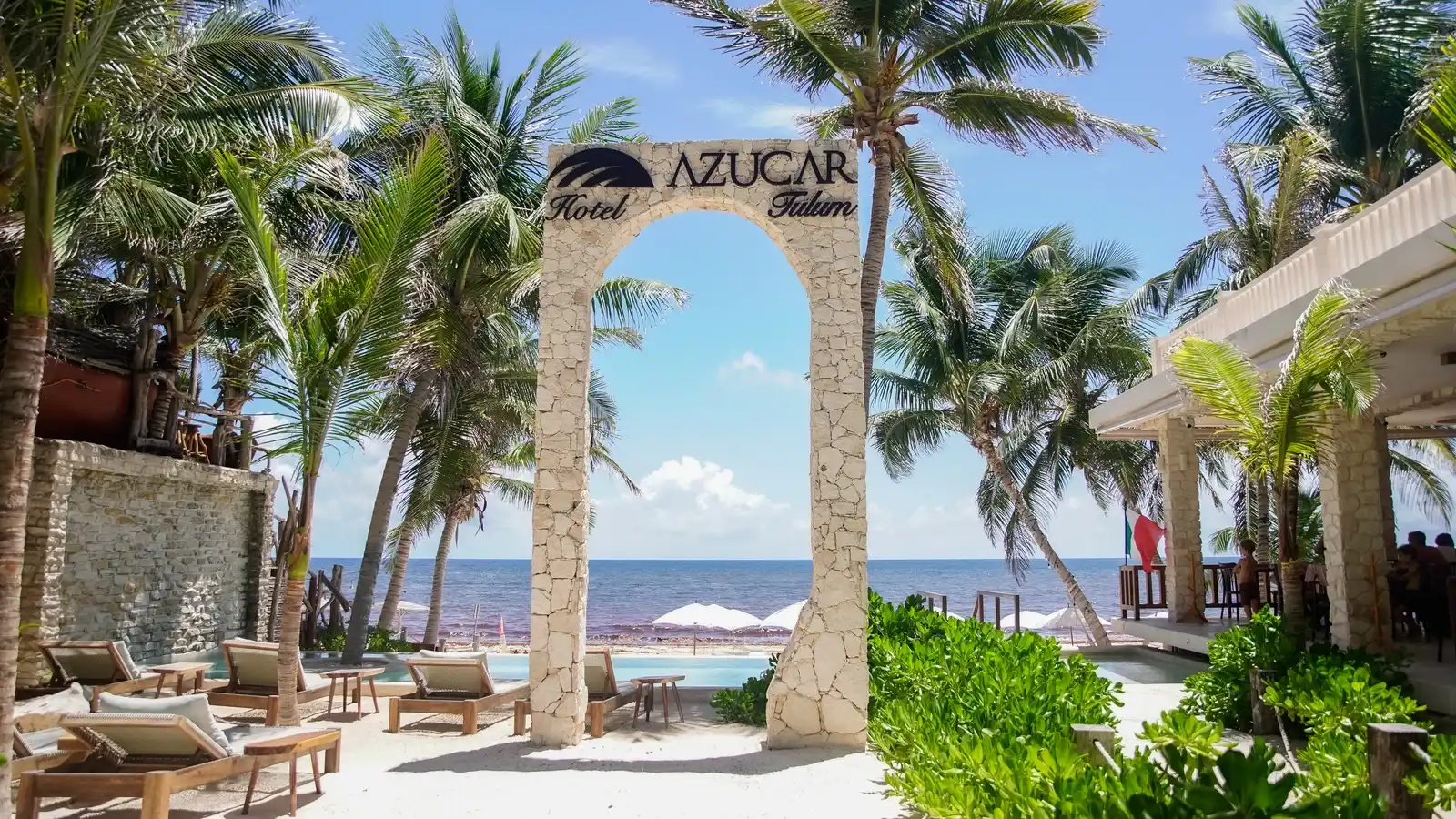 Arco de piedra con el rótulo del hotel, entre palmeras, con los camastros de la playa y el Caribe detrás.