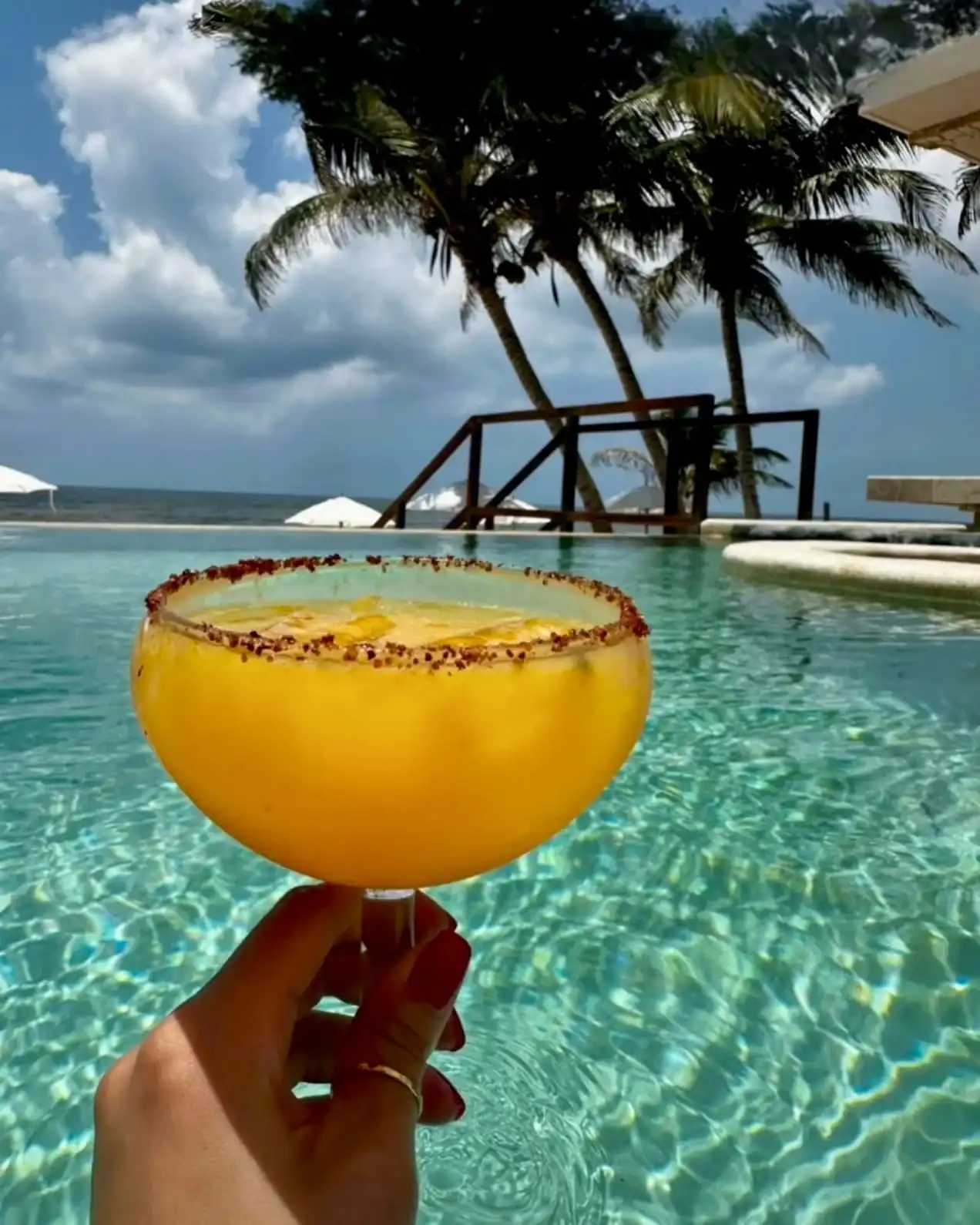 Una margarita de mango con el borde escarchado de chile, sostenida a contraluz frente a la alberca, con las palmeras y el mar al fondo.