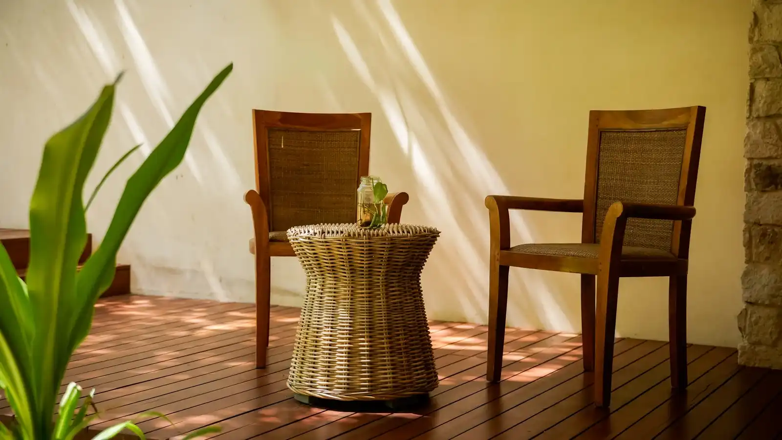 Dos sillas de madera y una mesa de mimbre en un entarimado, junto a un muro claro.
